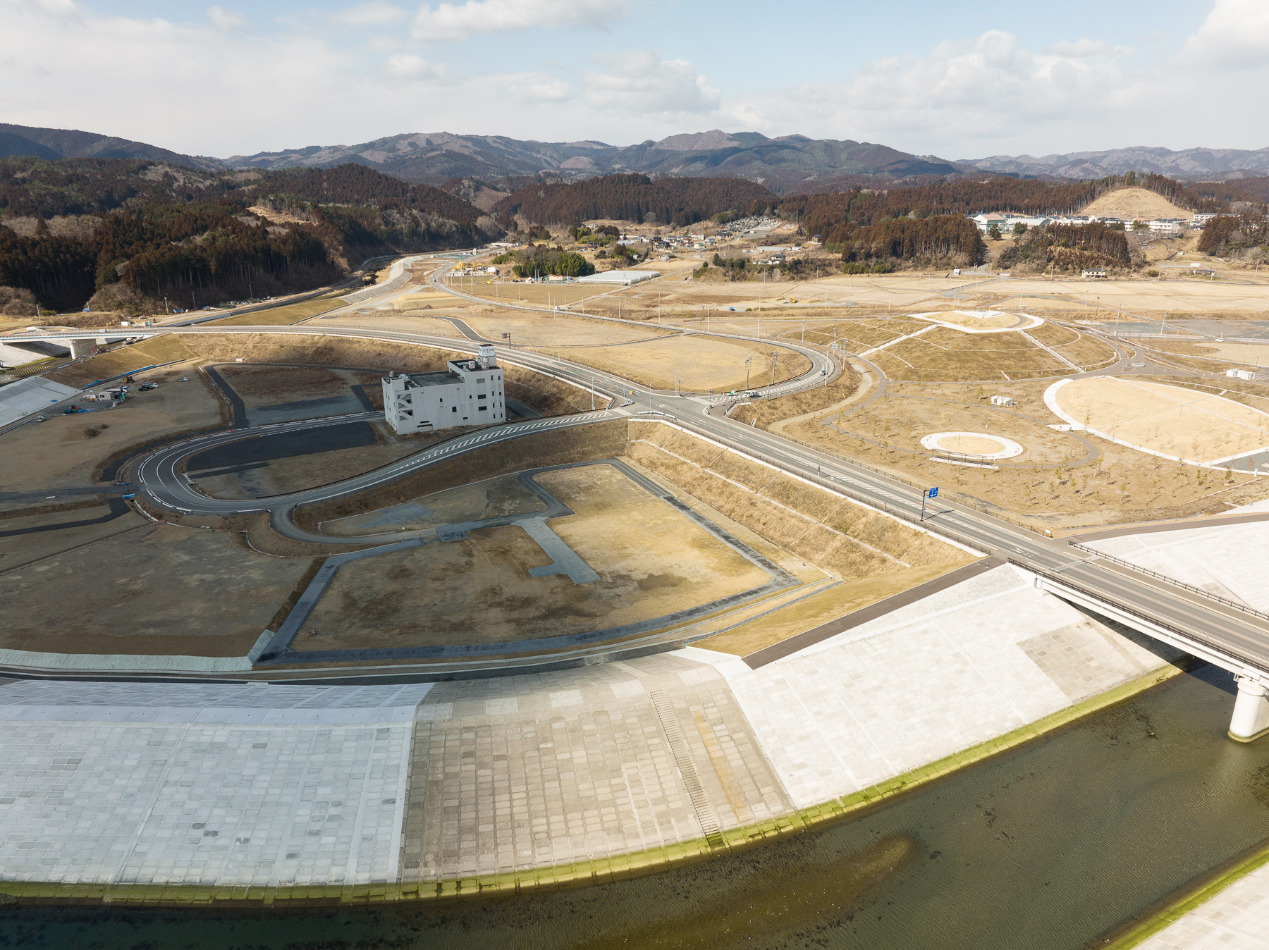 コンクリート護岸の内側に複雑に曲がる道路があり、その奥に山々と造成地が広がる津波復興地区の昼間の風景を高台から定点カメラで撮影した写真