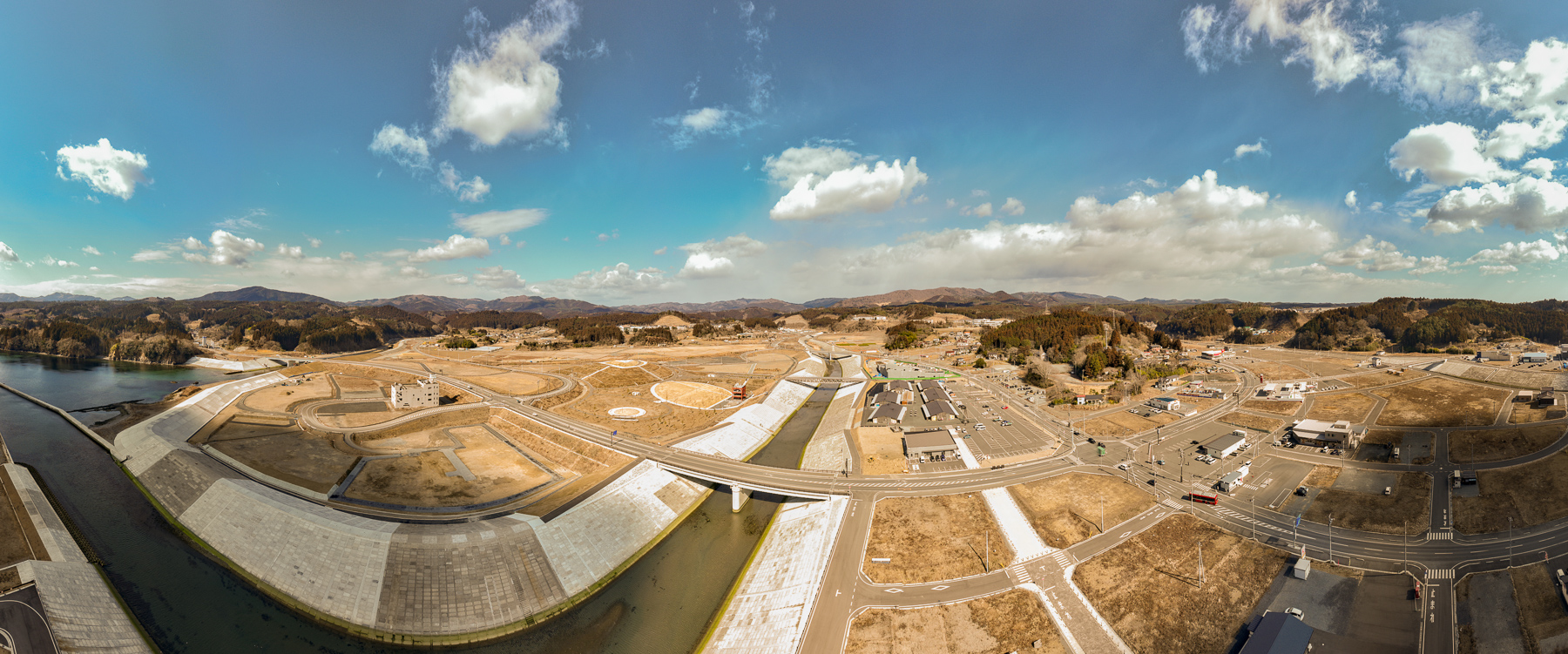 広い造成地に曲線状の道路が通り、中央に円形の盛土が複数あり、左に川、右に建物が点在する復興途中の街の昼間の風景を高台から定点カメラで撮影した写真