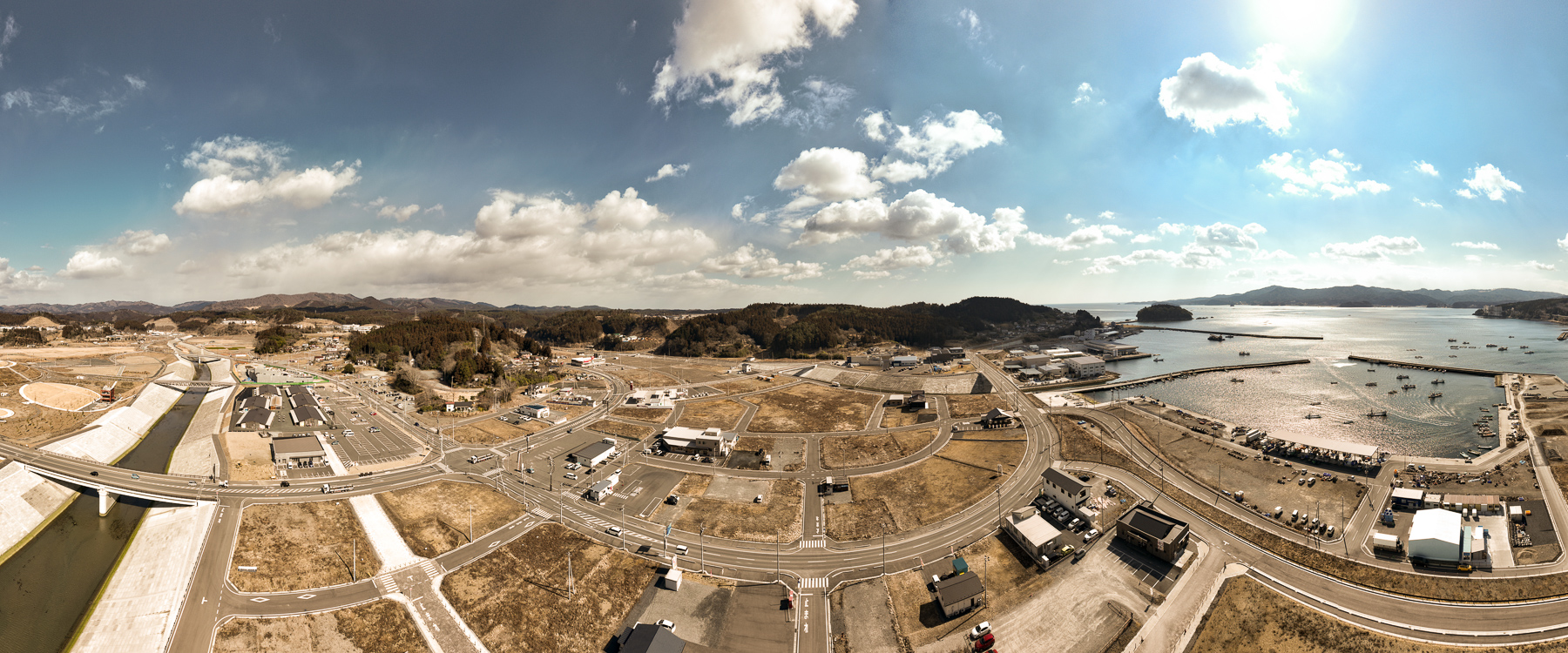 手前に港、奥に住宅や施設が並び、左には造成地と川、右には湾が見える復興港町の昼間の風景を高台から定点カメラで撮影した写真