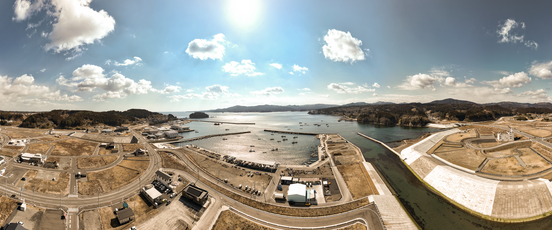 中央上に太陽が輝く空の下、湾内に多くの漁船が浮かび、右には護岸と造成地、左には漁業施設が見える沿岸都市の昼間の風景を高台から定点カメラで撮影した写真