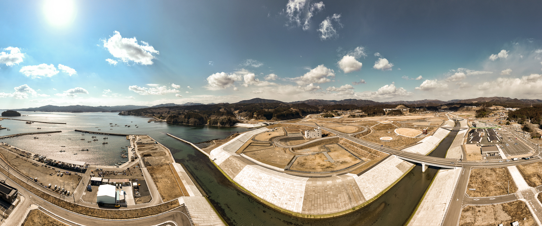 左に漁港と湾、右に広がる造成地と川が一直線に延び、中央に円形の造成跡と道路が幾重にも重なる復興中の街全景の昼間の風景を高台から定点カメラで撮影した写真