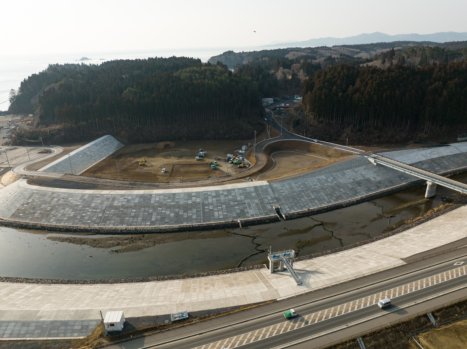 中央に広く平坦な造成地と建設機械が点在し、川を挟んで左右に高い護岸が整備され、背後に森と山が広がる昼間の風景を高台から定点カメラで撮影した写真