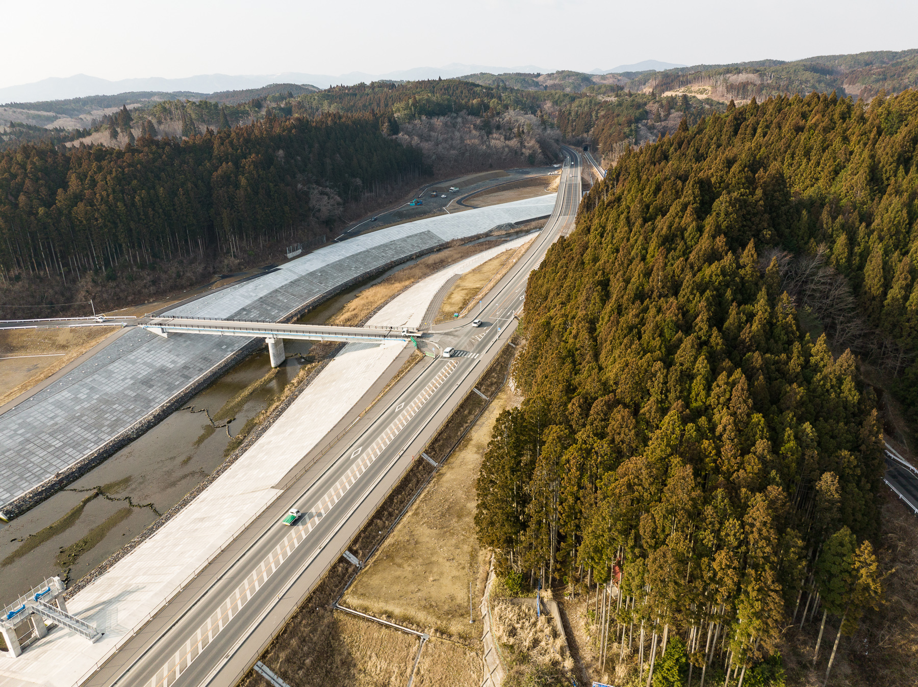 川沿いに続く防潮堤と車が走る道路が交差し、右手には広がる森林が見え、山あいに延びる新しい道が見える昼間の風景を高台から定点カメラで撮影した写真