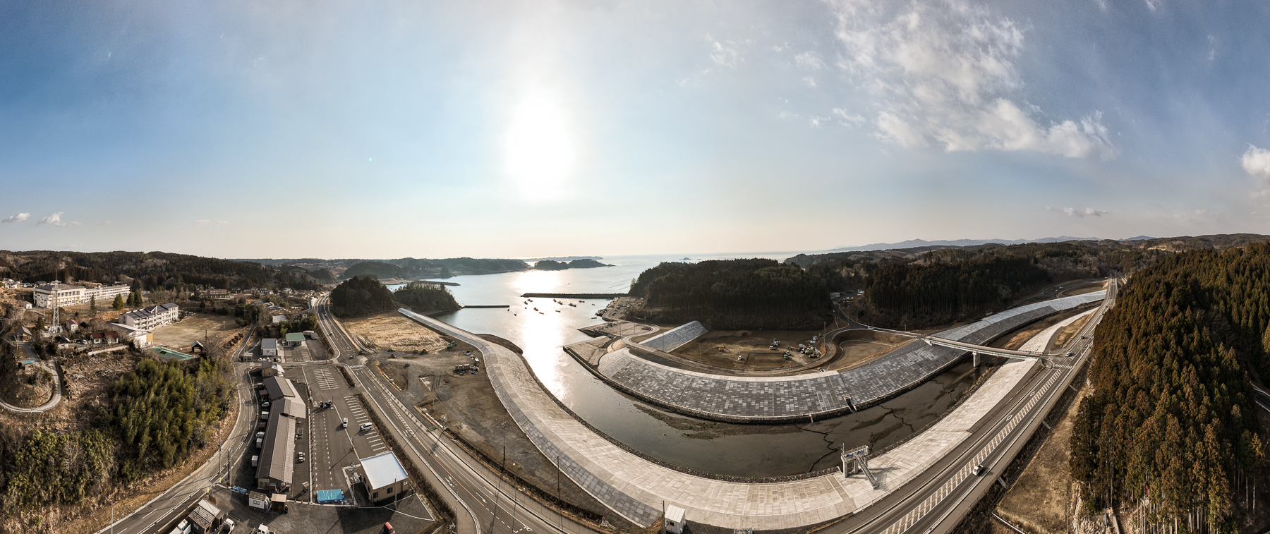 太陽が低く傾いた海辺の風景で、中央に蛇行した川が海に注ぎ、両岸には広大な護岸が広がり、道路や小さな集落が山に囲まれるように点在している昼間の風景を高台から定点カメラで撮影した写真