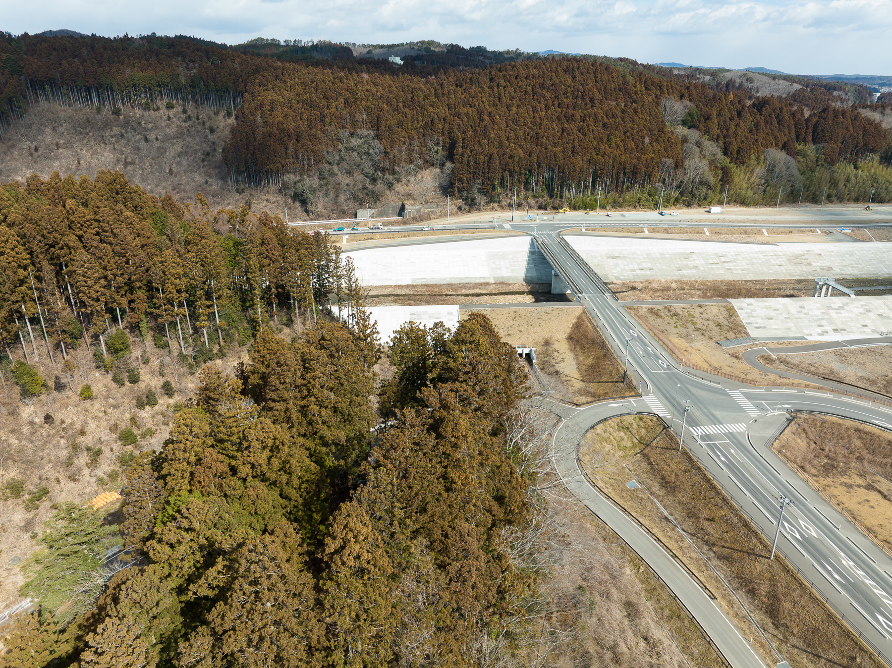 森林地帯のそばにある交差点と高架道路が写っており、周囲には舗装された広い空き地が広がる造成地の様子が分かる昼間の風景を高台から定点カメラで撮影した写真