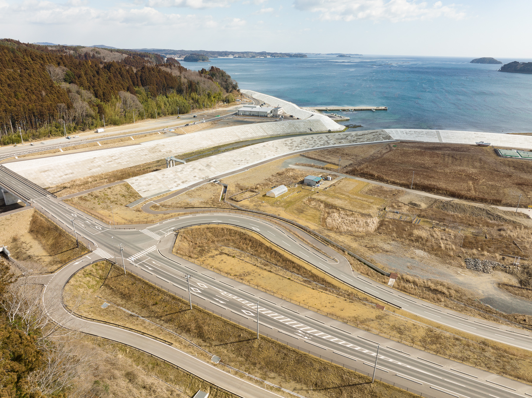 奥に広がる海と港の前に、防潮堤と見られる白い護岸や整地された土地が並び、道路と交差点が整備された沿岸の開発地域の昼間の風景を高台から定点カメラで撮影した写真