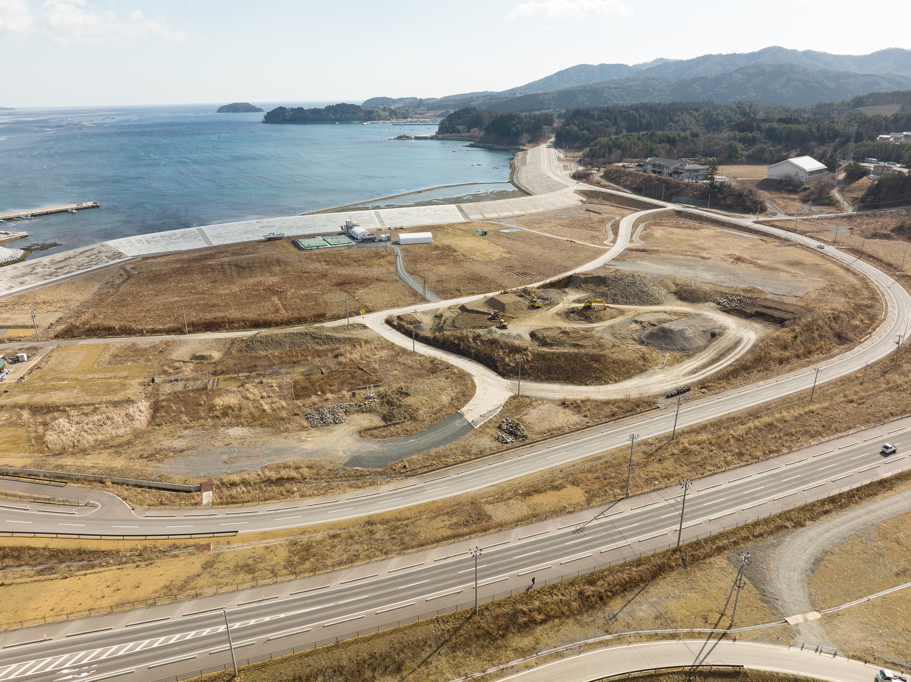 右奥にカーブしながら続く防潮堤と道路、中央には土を盛った造成地と建設機械があり、海と山に囲まれた復興地域の昼間の風景を高台から定点カメラで撮影した写真