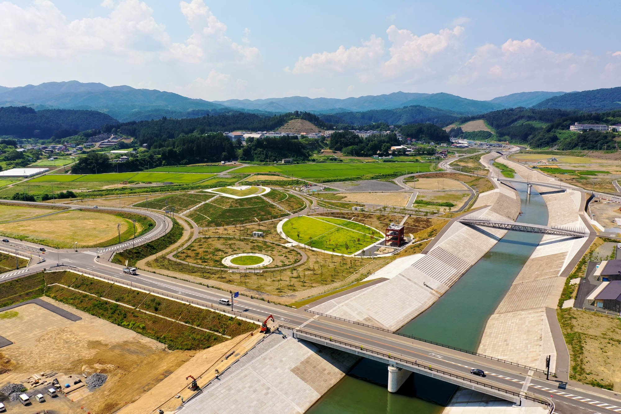 コンクリートで整備された川に橋が架かり、周囲には緑の草地と丘が広がり、草地には曲がりくねった道や円形の模様が見られ、遠くには建物やインフラが点在し、山々と雲が浮かぶ空を背景にした空中からの風景の写真
