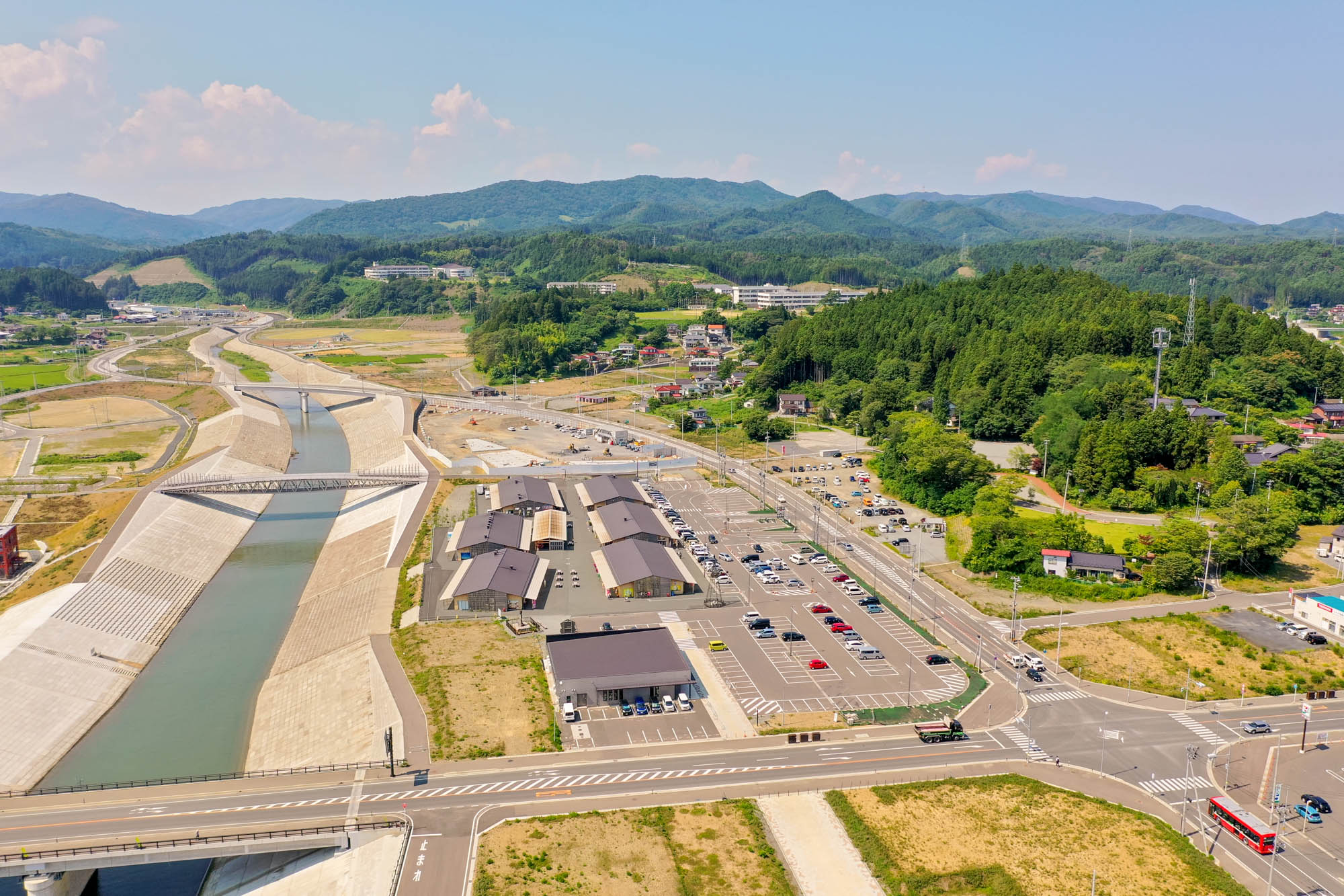 河川の両岸に新しく整備された護岸と広い道路が延び、建物と駐車場、奥に続く住宅地や緑豊かな山々が見える、志津川地区の復興が進む様子の写真