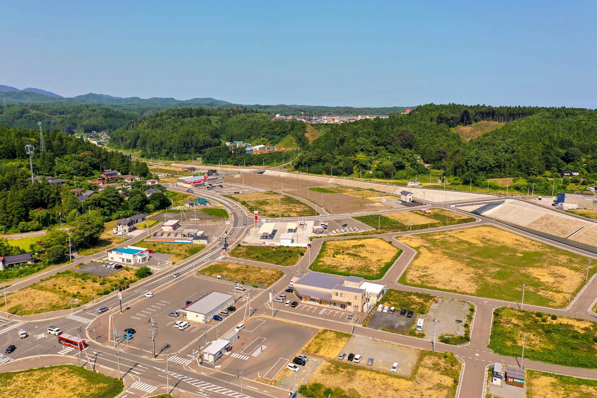 緑豊かな丘陵地帯を背景に、複数の道路が交差し、建物や駐車場が点在している広々とした開発地域の写真