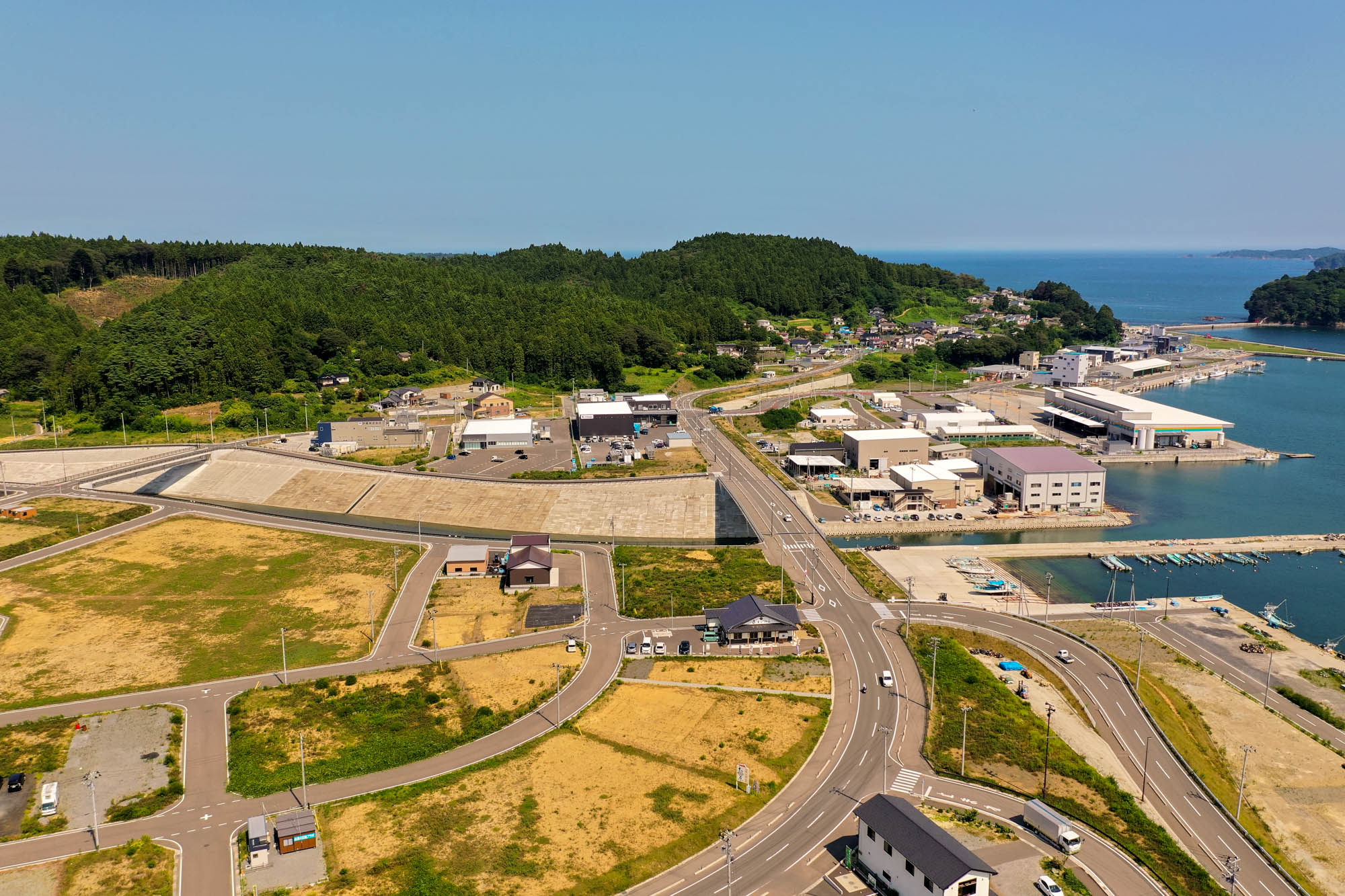 海に面した町を空から見下ろした風景で、建物や道路が整然と並び、港には複数の桟橋が海に突き出し、町の周囲には緑や丘が広がり、いくつかの区画には建物が建ち、他は空き地や開発中の様子が見られる海辺の町の写真