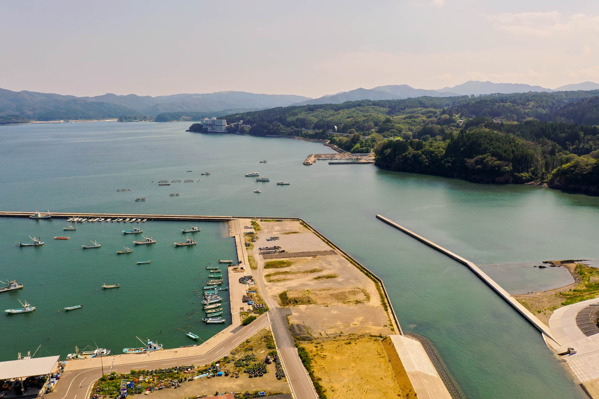複数の船が停泊する静かな海の港を中心に、周囲には道路や建物が整備され、背景には緑の丘と山々が広がり、防波堤が海に突き出して港を守っている海岸地域の写真