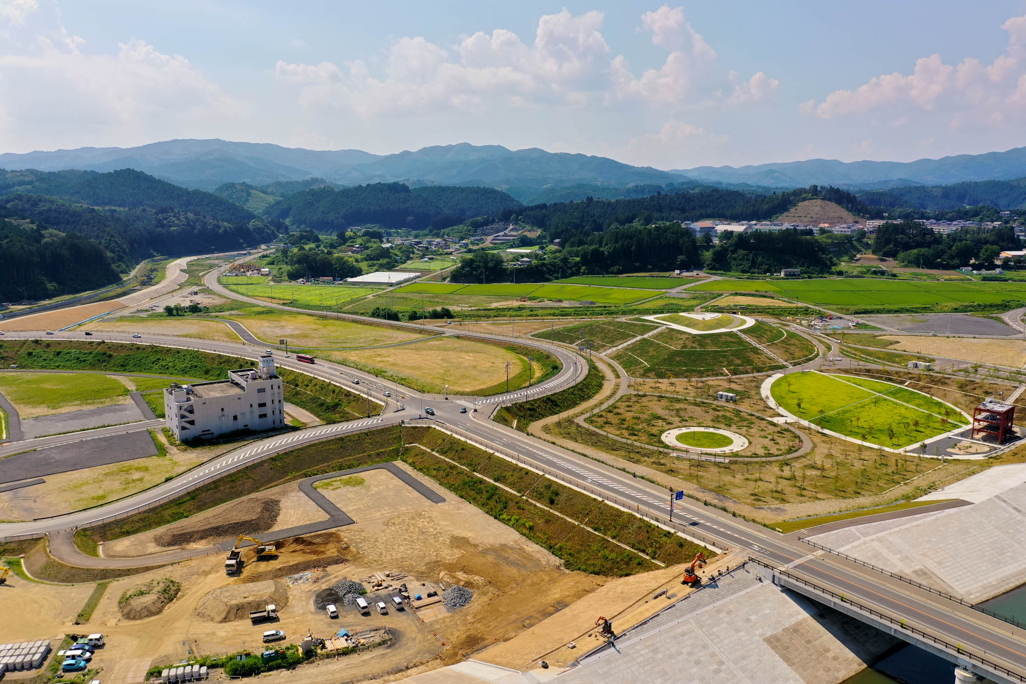 緑の田畑と建設中の区画が広がる風景の中に道路網が走り、背景には山々が連なり、ところどころに建物が点在し、晴れた空に雲が浮かぶ開発地域の写真