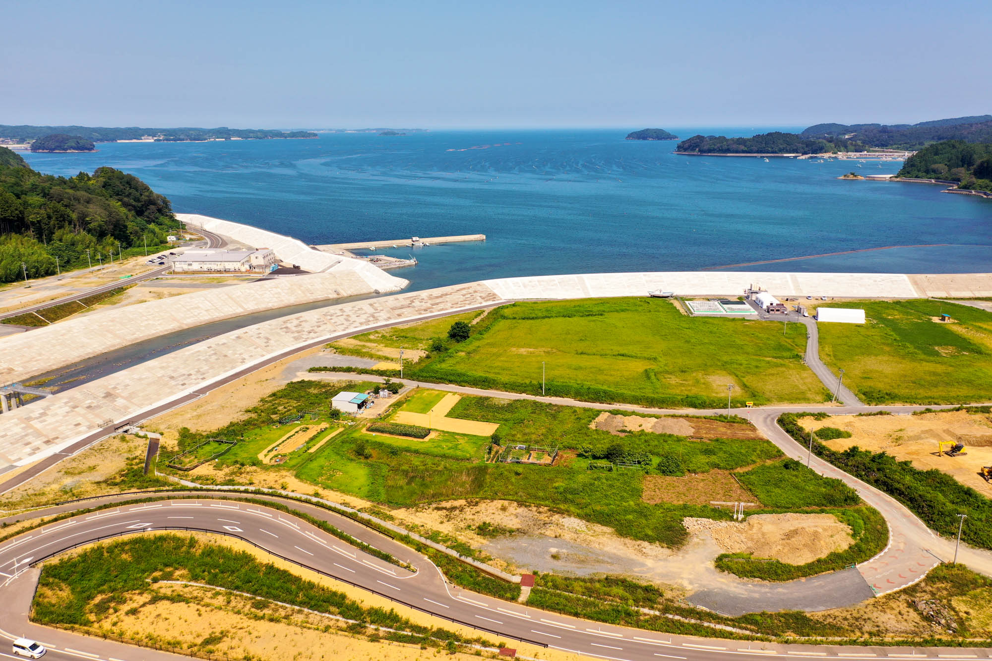 手前に湾曲した道路が走り、その周囲には緑の畑が広がり、奥には海が見える沿岸地域を空から見た写真