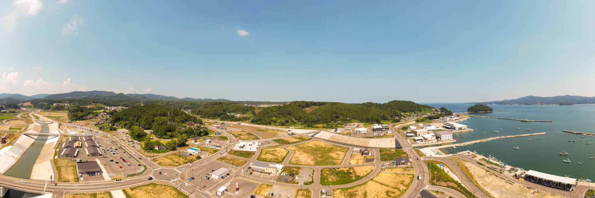 右側に船が停泊する桟橋のある海が広がり、町にはさまざまな建物や道路、緑地が点在し、背景には丘や森林が広がっていて、澄んだ青空の下に広がる海辺の町のパノラマ風景の写真