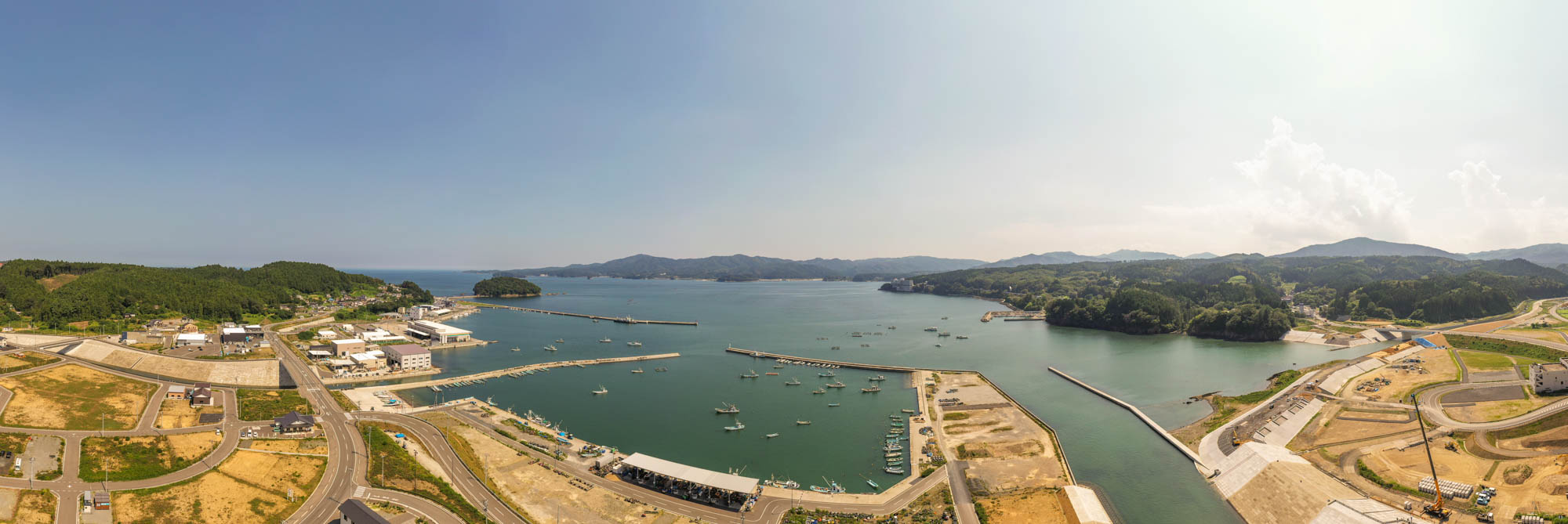 港に複数の船が停泊している海辺の地域を広く見渡す風景で、周囲には建物や道路、緑の丘が広がり、遠くには雲が浮かぶ澄んだ空が見える海岸地域の写真
