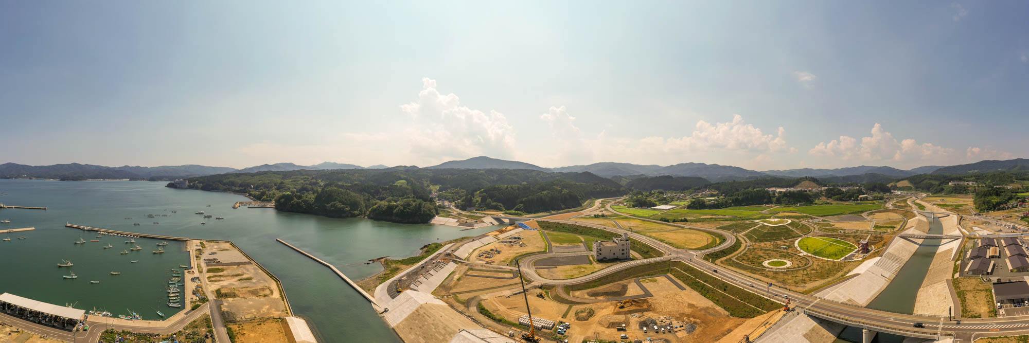 左側に複数の船が停泊する港があり、海岸線にはコンクリートの構造物や桟橋が並び、右側には道路や通路、建物が見える広い造成地が広がり、背景には丘や山々が連なり、晴れた空に雲が浮かぶ海岸地域の写真