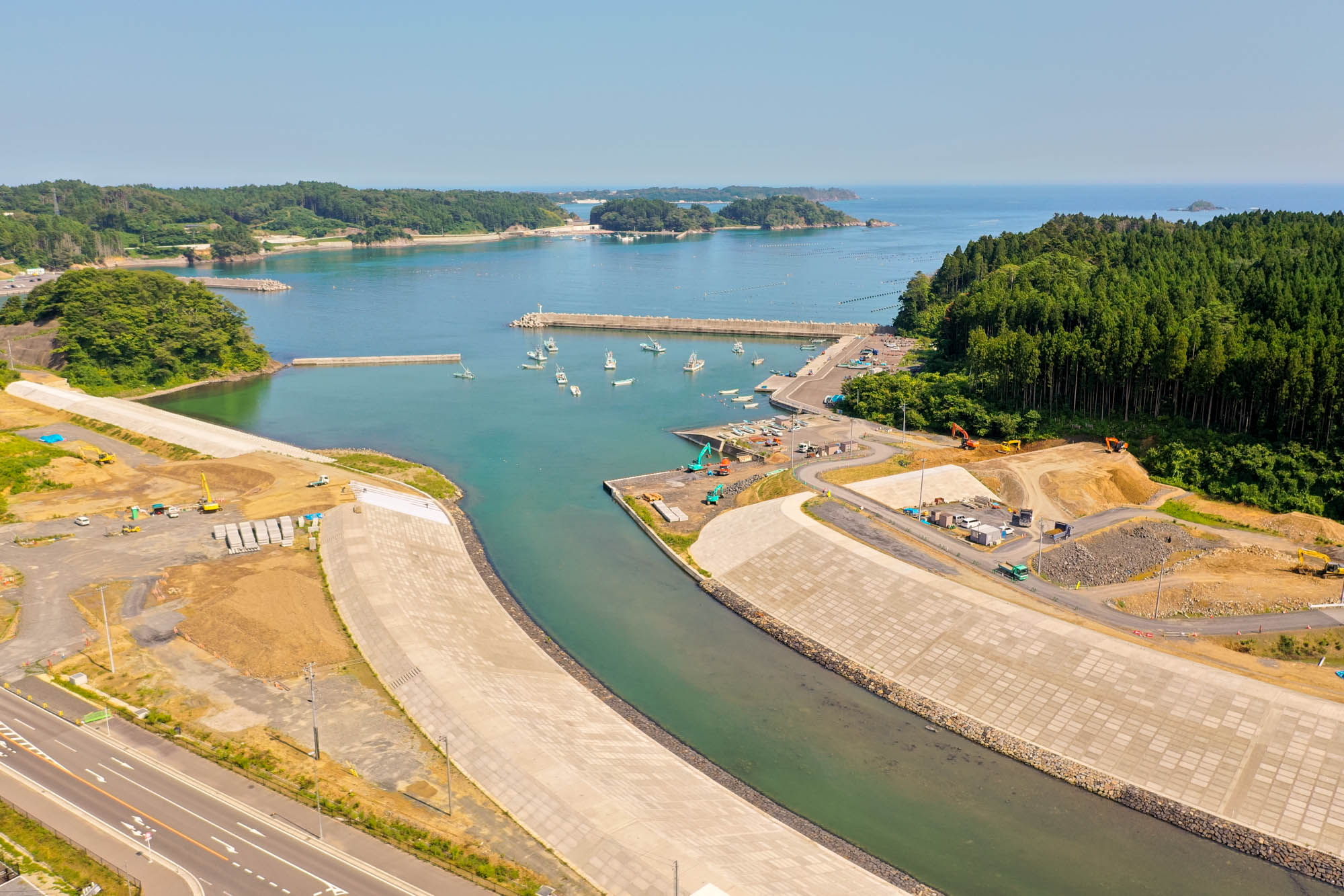 複数の船が防波堤に囲まれた港に停泊し、周囲には森林や空き地、建設現場が広がり、左下には道路が走り、海岸沿いにはさまざまな構造物や機材が見える海辺の地域の写真