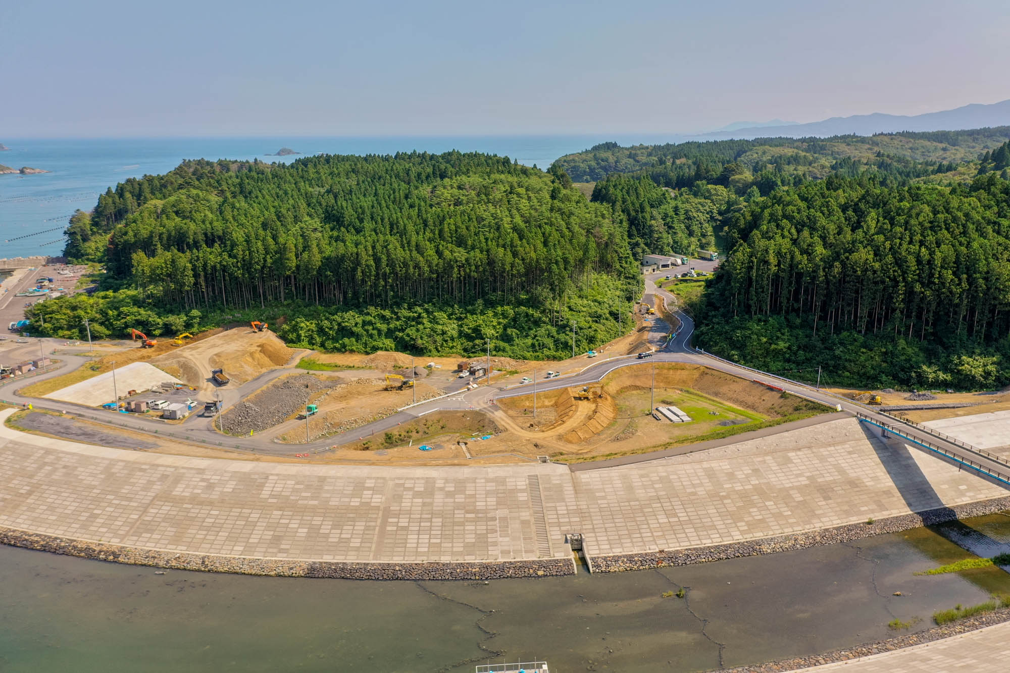 手前に大きなコンクリートの防波堤があり、その背後には重機や車両が並ぶ工事現場が広がり、両側には森林が生い茂り、奥には海と小さな島々が見え、青空の下に広がる海岸地域の写真