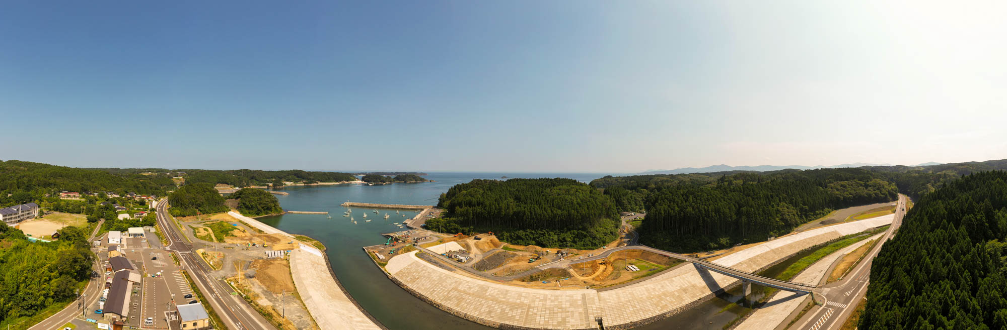 森林に覆われた丘に囲まれた湾のある海岸地域を広く見渡す風景で、水辺には防波堤のような構造物があり、道路やインフラも整備され、晴れ渡った青空の下に広がる海辺の写真