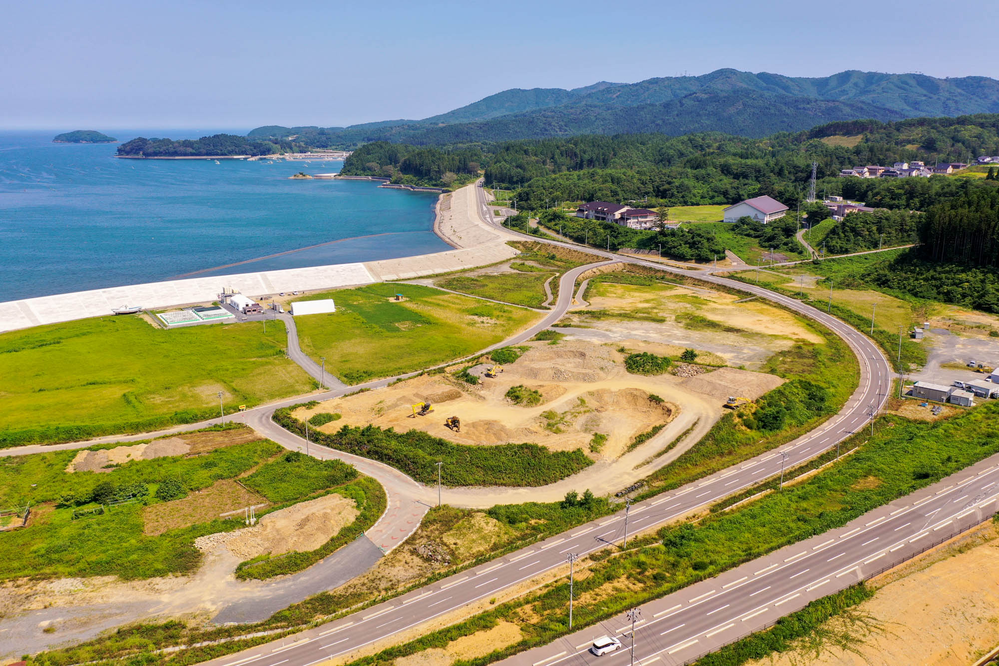 左側に海と防波堤が広がり、手前には曲がりくねった道路と緑の畑、工事や掘削現場が点在し、背景には森林に覆われた丘と建物が散在する、青空の下の沿岸地域の写真