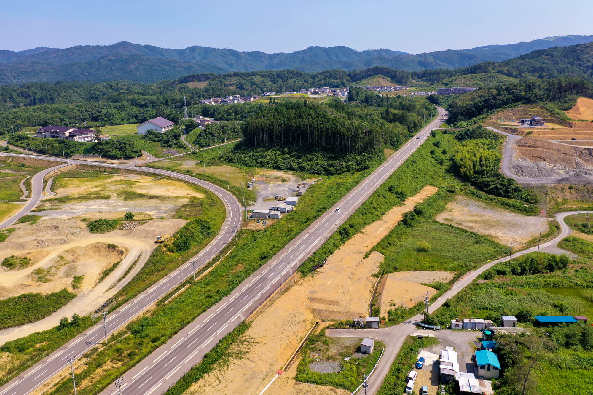 高速道路が緑の丘陵地帯を貫いて走り、周囲には工事現場や建物、住宅が点在し、背景には木々に覆われた山々が広がる戸倉地区の風景を空から見た写真