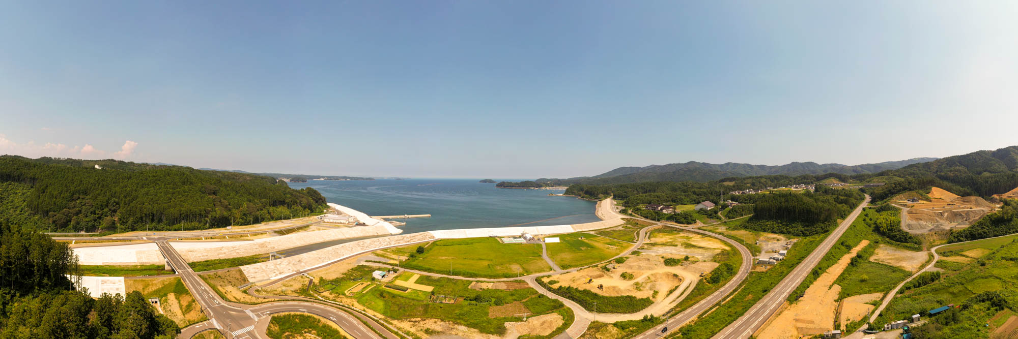 広大な海を囲むように緑の丘や森林が広がり、海岸線には道路や開発中と思われる土地が点在し、雲ひとつない青空の下に広がる沿岸地域の写真