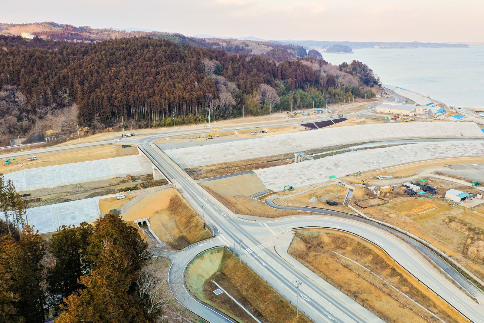 複数の道路が交差しながら曲がりくねって広がる地域を上空から撮影したもので、背景には森林に覆われた丘が連なり、遠くには海が見え、道路周辺の土地は開発中または工事中の様子で、整地された場所や点在する構造物が確認できる冬の海沿いの造成地の写真