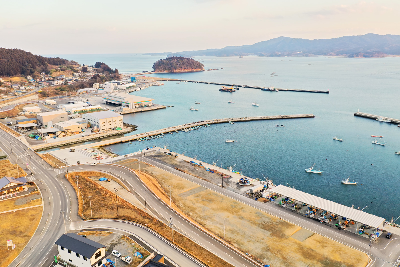 港には複数の船が停泊し、海岸沿いにはさまざまな建物や構造物が並び、周囲には道路と緑地が広がり、背景には丘が連なり、海の向こうには島が見える海辺の町を写した冬の港町の写真