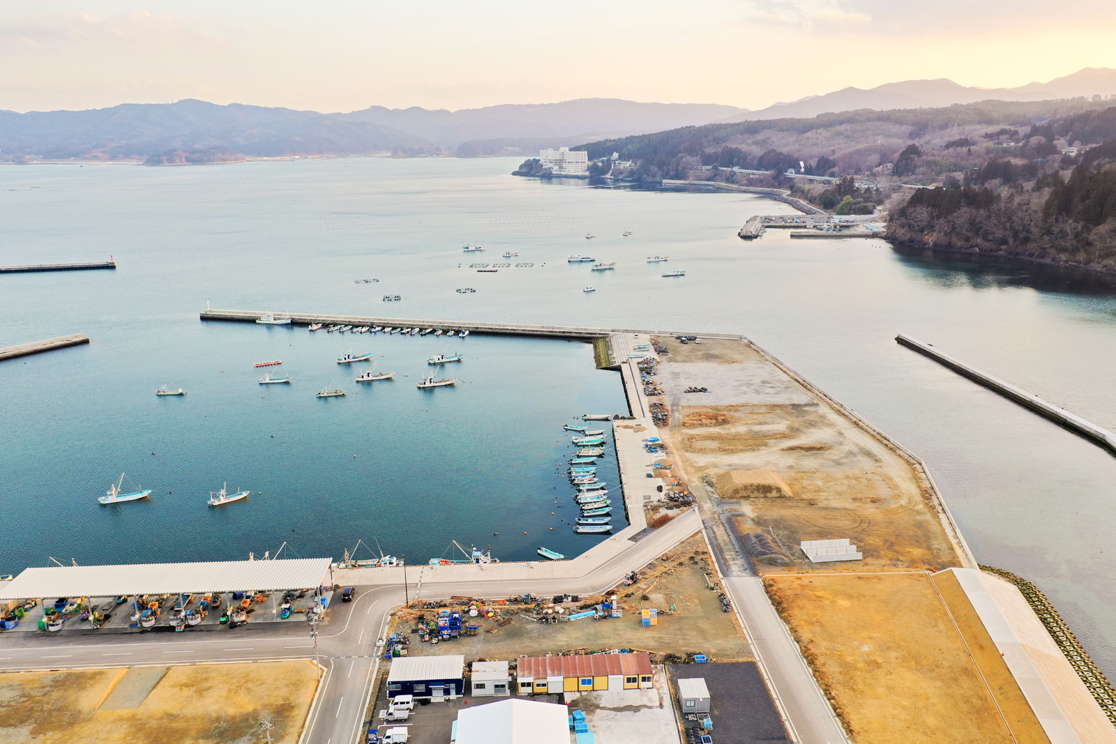 港には複数の船が停泊し、周囲には建物や道路、空き地が広がり、海岸線は遠くまで続き、地平線には丘や山が連なる海沿いの町を上空から撮影した冬の港町の写真