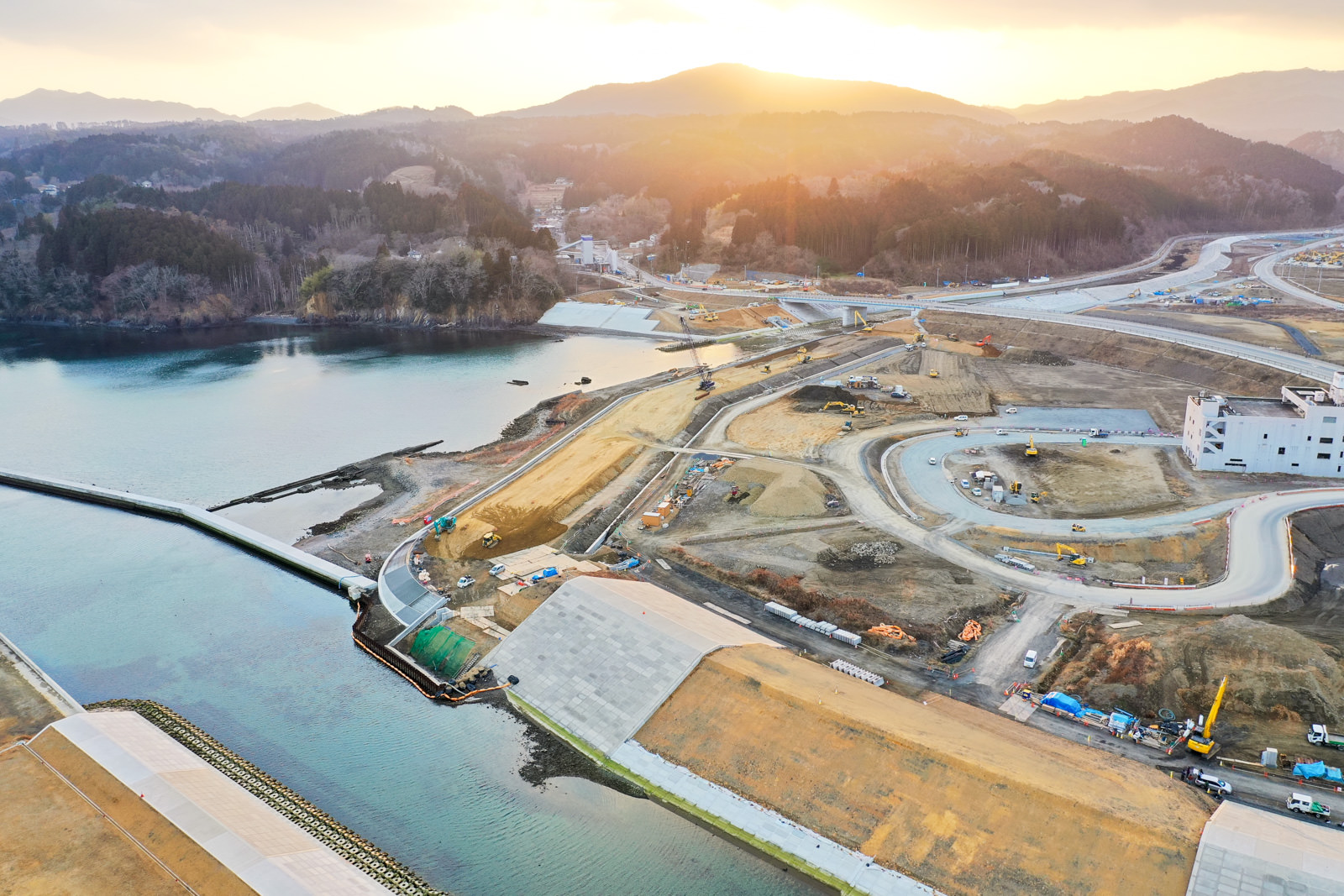 湾または川の近くに位置する沿岸の建設現場が写っており、複数の曲がりくねった道路、建設車両、建設中の建物が点在していて、背景には山々が見え、太陽が昇るか沈むかしている時間帯で、全体に暖かい光が差し込んでいる写真