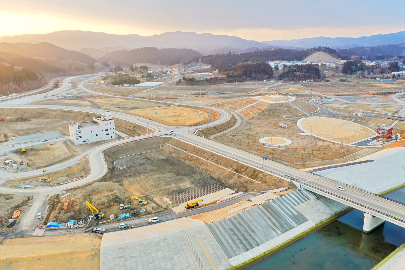 広大な建設現場が写っており、複数の道路や橋、クレーンやトラックなどの建設車両や機材が点在していて、背景には山々が広がり、周囲の風景は草地や土が混在するほぼ荒れ地で、右下には川または運河が流れ、それに架かる橋も見える写真