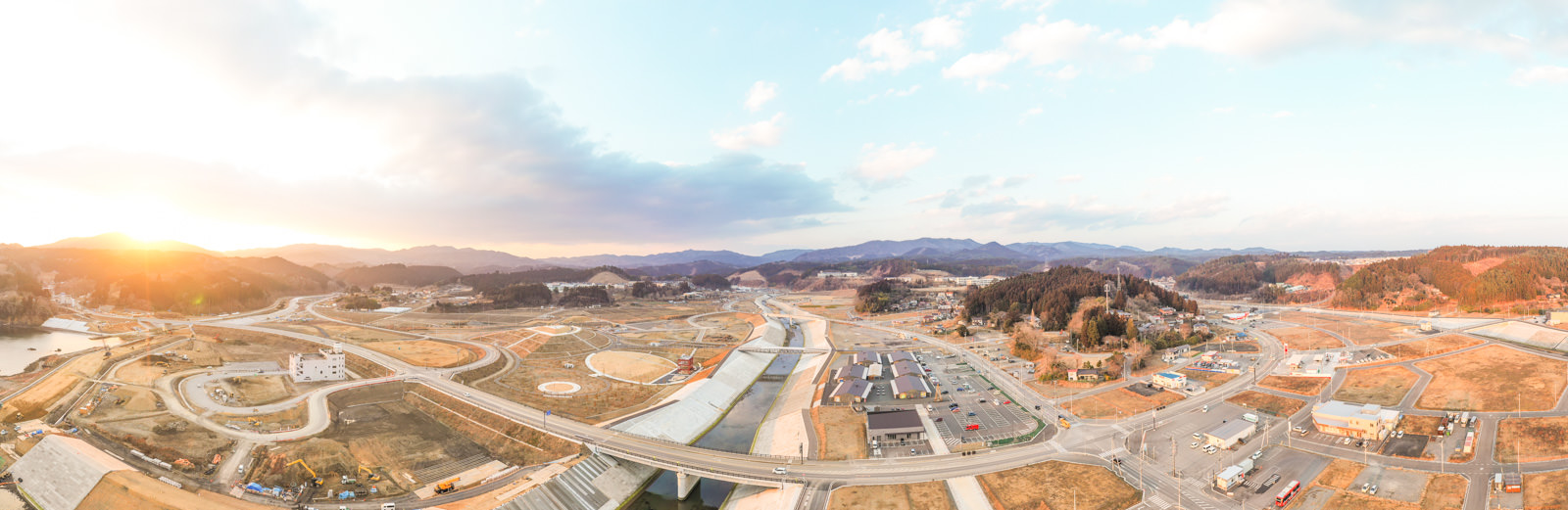 道路や橋、建物などのインフラが点在する風景を広く見渡す構図で、手前には開発された土地と自然のままの土地が混在し、背景には山々が連なり、太陽が地平線近くに位置する日の出または日の入りの時間帯に撮影された冬の山間の町の写真