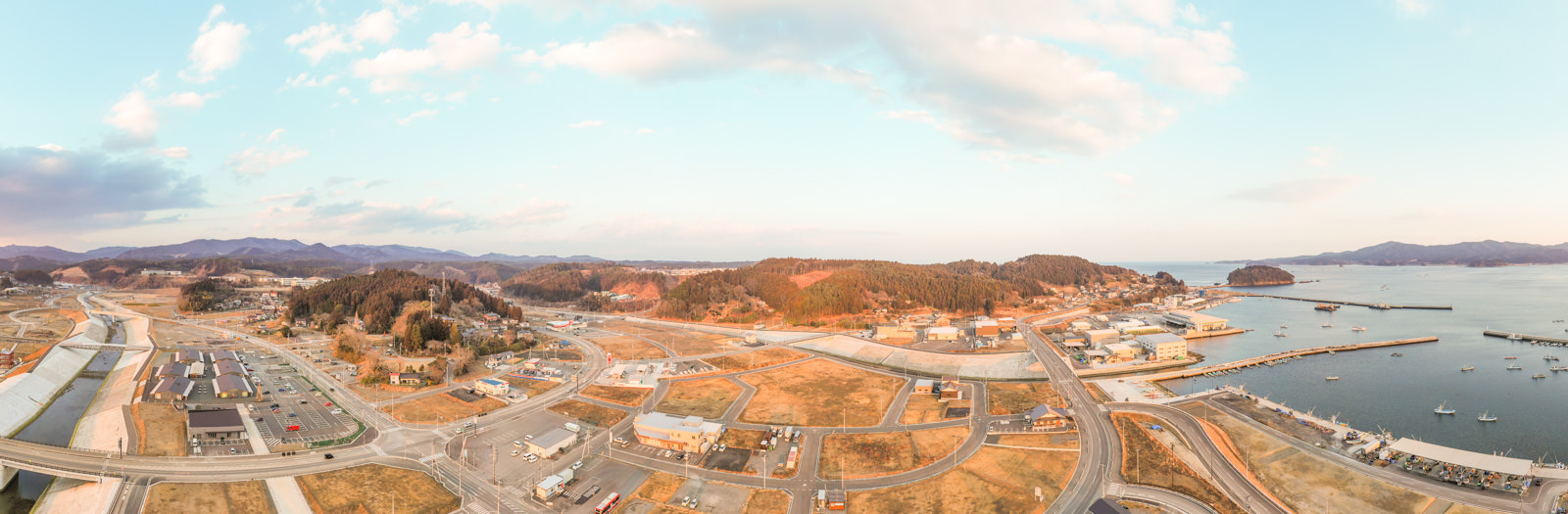 海沿いの町を上空から撮影したもので、右側に海が広がり、町にはさまざまな建物や道路、港があり、背景には丘や山が連なり、空は淡い青色で雲が点在する冬の海辺の町の写真
