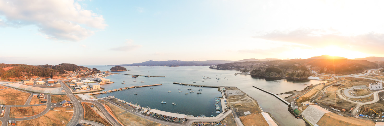 港には複数の桟橋と船があり、町は海岸線に沿って広がり、建物や道路が入り組み、背景には丘や山が連なり、右側には太陽が昇るまたは沈む様子が見られ、暖かい光が風景を照らしている冬の海辺の港町の写真