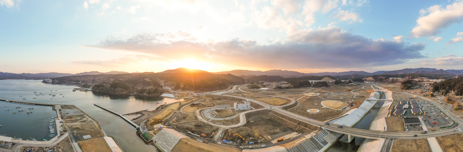 広い湾が穏やかに広がり、その周囲には建設途中の建物や整備中の土地が点在しており、空には夕日が赤く染まりながら沈みかけていて、全体が静かで広々とした雰囲気に包まれている写真