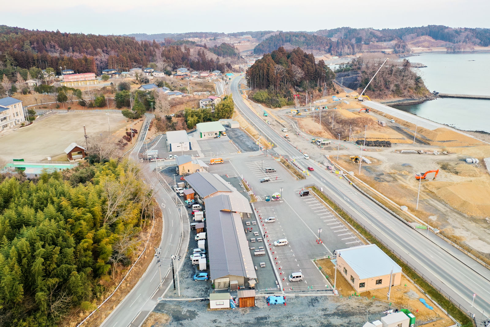 海岸沿いの地域を上空から見下ろした風景が広がっており、道路が地域を縦断し、複数の建物や駐車場、車両が点在していて、森林地帯や開けた草地、海岸近くの建設現場が混在し、海岸線には小さな湾または入り江があり、そこにも建設機材が見える写真