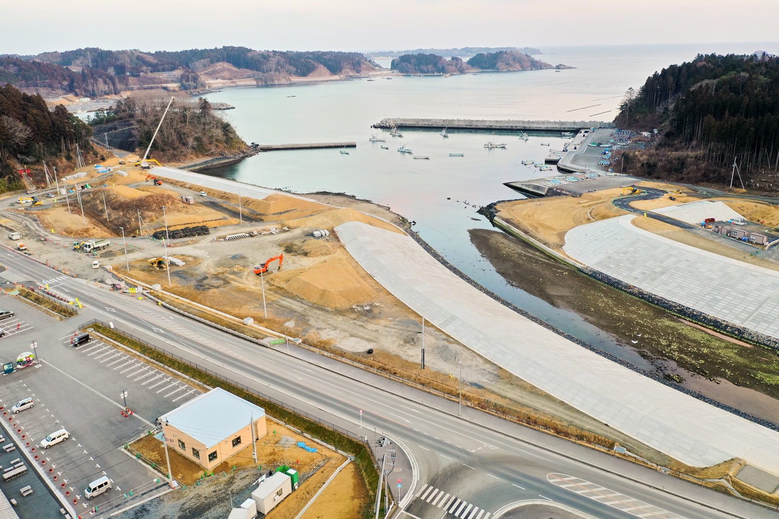 開発または建設中の海岸地域を上空から見下ろした風景が広がっており、手前には道路や駐車場、建物が点在し、中ほどには建設機材や資材が並び、海岸沿いには大きなコンクリート構造物が建設されていて、背景には桟橋に停泊する船とそれを囲む森林に覆われた丘が見える写真