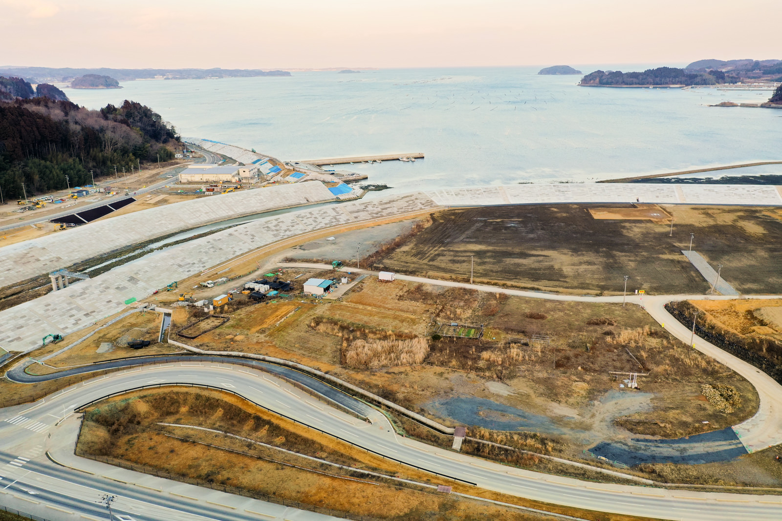 手前に曲がりくねった道路が伸び、その先には平坦で開けた土地が広がり、点在する小さな建物や構造物が見られ、背景には海が広がり、水平線上には複数の小さな島々が浮かび、全体的に自然が多く残る開発の進んでいない海沿いの地域を写した冬の沿岸風景の写真