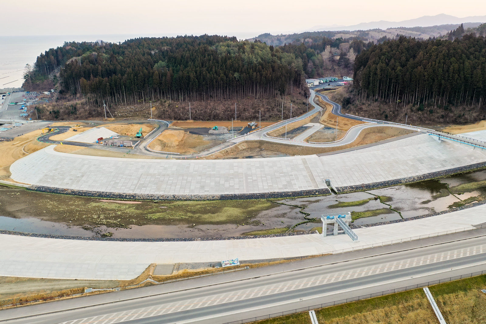 広大なコンクリート製の治水構造物（おそらく堤防）が上空から写されており、手前にはそれに沿って走る道路があり、構造物は部分的に乾いた川床または排水区域を囲むように湾曲していて、周囲には複数のアクセス道路や通路、小規模な建物や建設機材が点在し、背景には森林に覆われた丘や山々が広がっている写真
