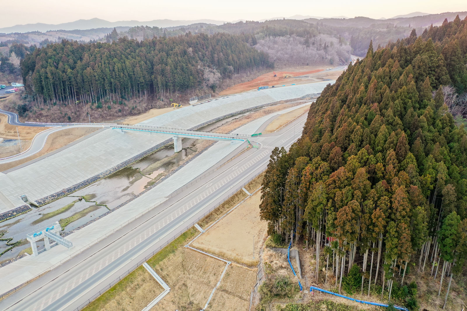 広大な風景の中に新しく建設されたコンクリート構造物があり、橋がその上を横断していて、右側には背の高い木々が密集した森林が広がり、背景には樹木や草木に覆われたなだらかな丘陵地が続いている写真