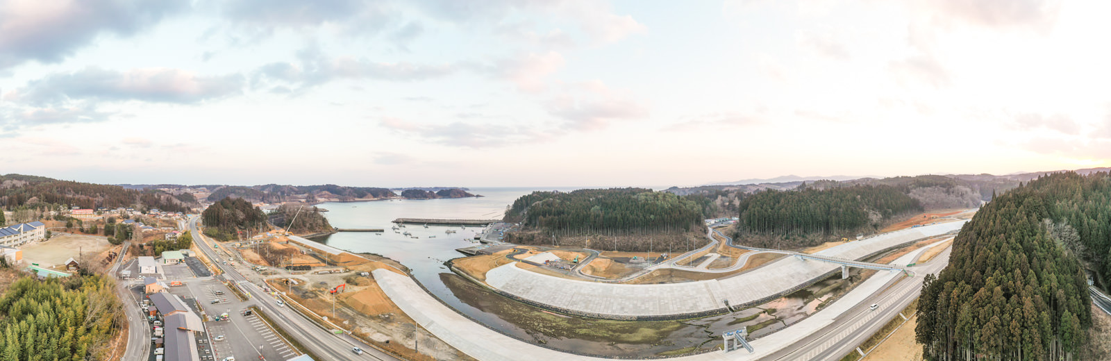 海または大きな水域を背景に、陸地が水面に向かって突き出している沿岸地域のパノラマ風景が広がっており、手前には道路や建物、橋や堤防などの人工構造物が点在し、特に右側には木々が密集した森林地帯が広がっている写真