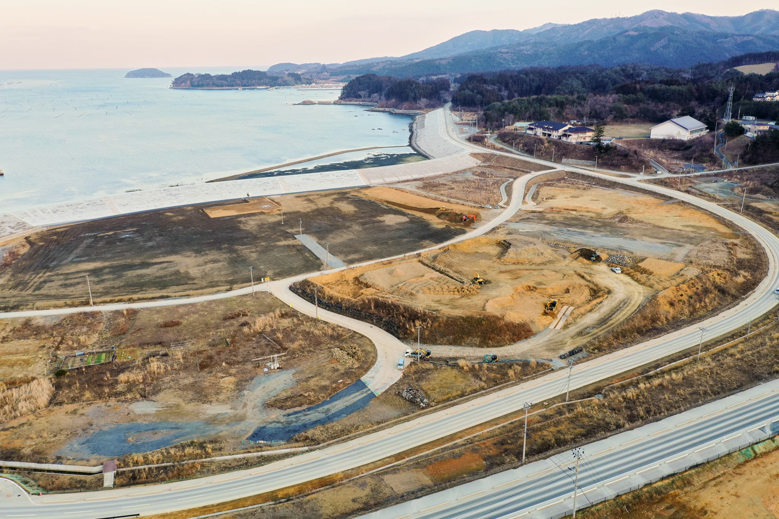 海沿いの地域を上空から撮影したもので、左側に海が広がり、曲がりくねった道路が通る周囲には整地された土地があり、開発中と思われる様子が見られ、背景には山々が連なり、植生はまばらで、遠くにいくつかの建物が点在する冬の沿岸部の造成地の写真
