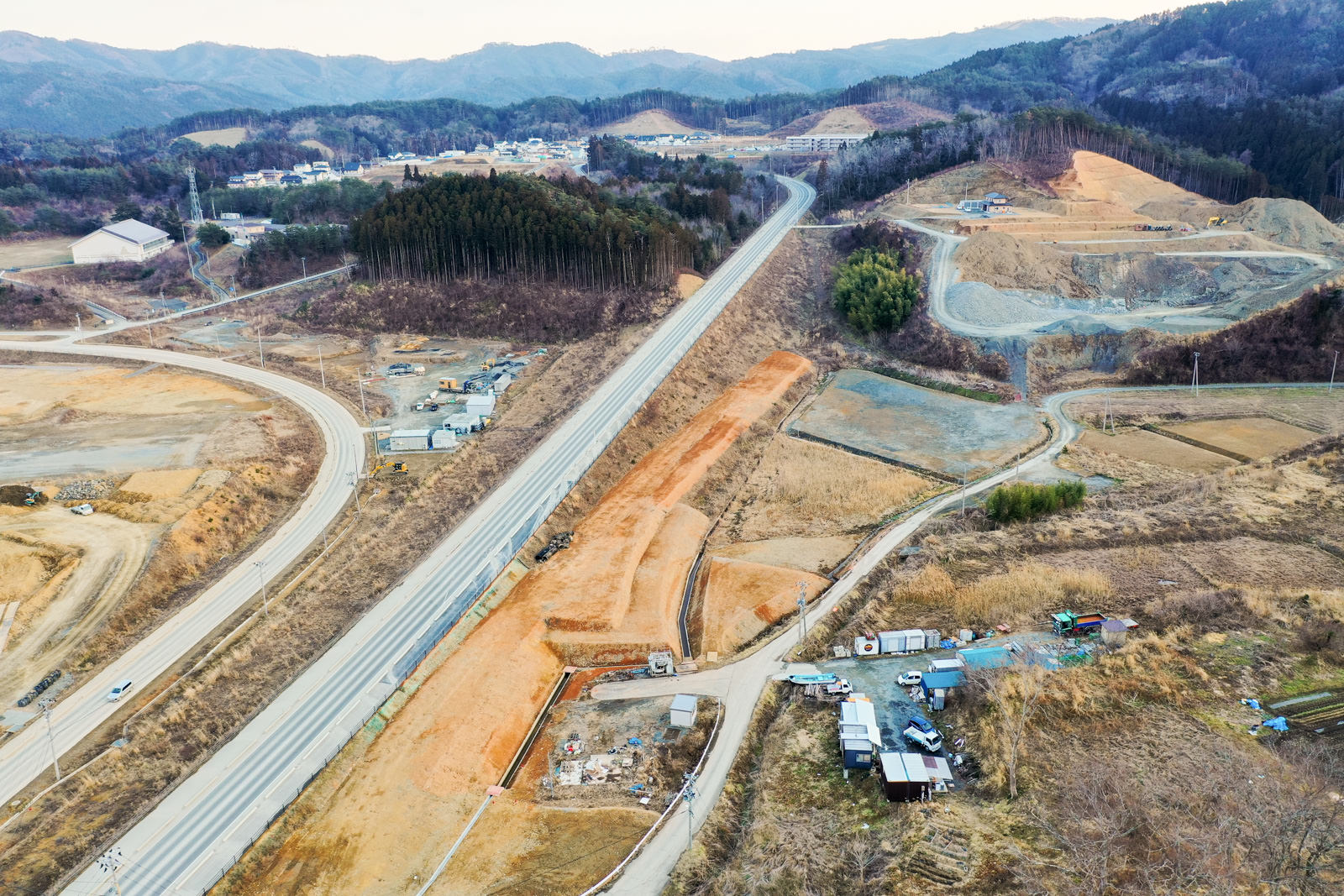 中央に高速道路が通り、その周囲には丘陵地と複数の建設現場が広がり、建物や構造物が点在し、整地された開発予定地が見られ、背景には山々と密集した森林が広がる冬の山間部の造成地の写真