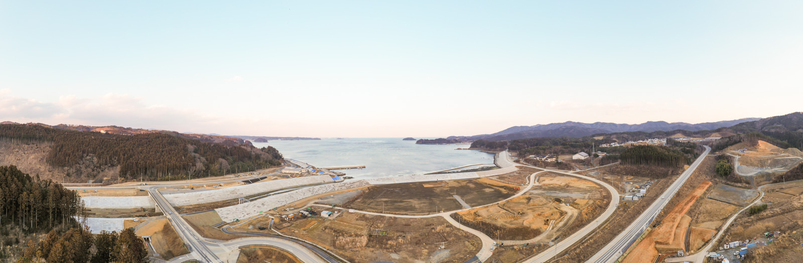 海沿いの町で、防潮堤が建設された湾と整地された広大な土地、曲線を描いて走る新しい道路と山々に囲まれた集落が遠くに見える写真