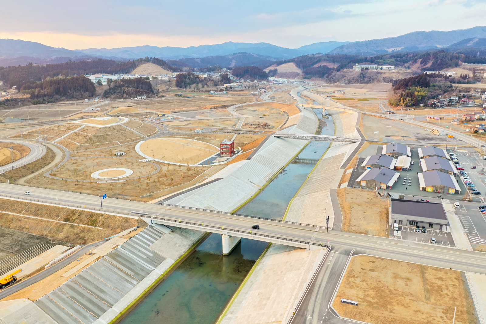 中央を川が流れ、その上に橋が架かり、両岸には道路や小道が広がり、右側には建物や駐車場が点在し、周囲には開発中または新しく整備された土地が見られ、背景には丘や山が連なる農村または郊外の風景を上空から撮影した冬の川沿いの町の写真