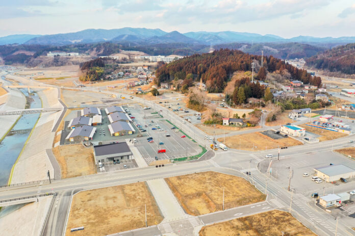 山に囲まれた町の一角で、整備された川とその横にある新しい商業施設と広い駐車場、背後には住宅地と林に囲まれた写真