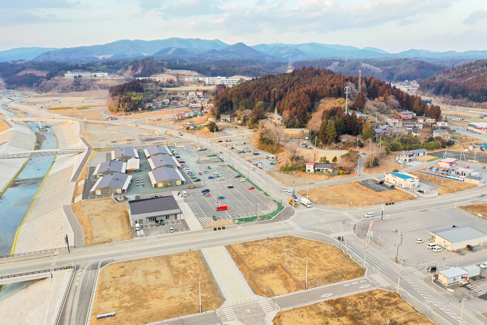 複数の建物や道路、駐車場が広がる場所を上空から撮影したもので、左側には川が流れ、中央右には木々に覆われた丘があり、背景には山々が連なり、空には雲が点在する写真