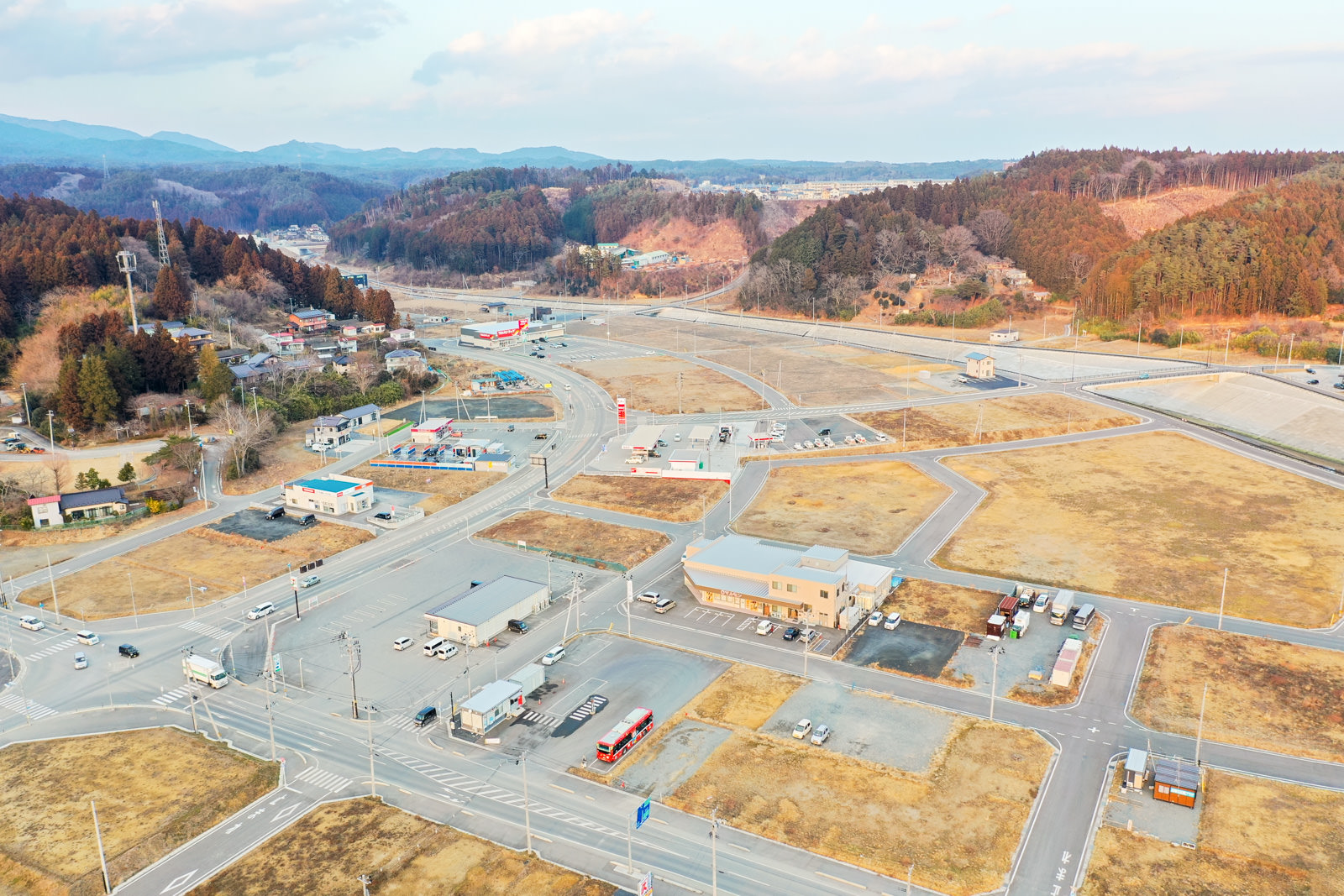 複数の建物や道路、広い空き地が広がる農村地域を上空から撮影したもので、住宅と商業施設が混在し、道路には車両が走っており、背景には森林に覆われた丘や山が連なり、空には雲が点在する冬の地方の町の写真