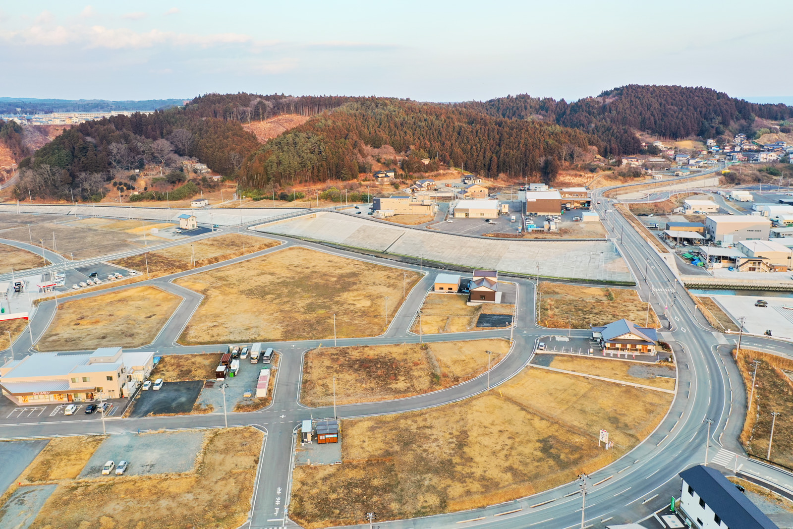 開けた畑や道路、建物が混在する地域を上空から撮影したもので、複数の区画に構造物が点在し、曲がりくねった道路がそれらを囲むように走り、背景には森林に覆われた丘と点在する建物が広がり、空は晴れて雲が少し浮かぶ町の写真
